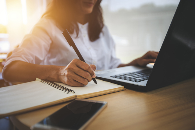 Person sitting in front of a laptop and writing on a piece of paper next to the laptop.